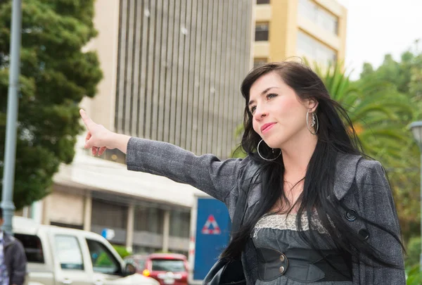 Classy latina model wearing smart casual clothes walking in urban street holding arm out signalling for taxi — Stok fotoğraf