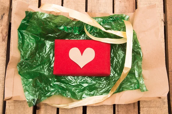 Nicely wrapped presents in red wrapping with golden heart lying on green plastic and brown paper, wooden surface background — Stockfoto