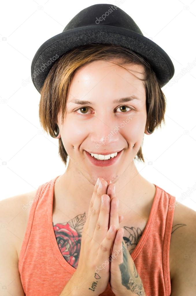 Headshot young hispanic male wearing black hat, red sleveless shirt with tattoos on chest and arms, punk rock insipired look, posing for camera folding hands as praying
