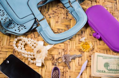 Womans purse light blue color lying flat with accessories such as mobile, makeup, keys and money spread out in front