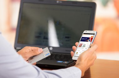Closeup of young mans hands holding smartphone up with Alibaba website visible, Visa card in other hand with laptop computer sitting on desk