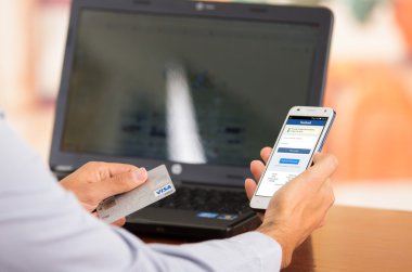 Closeup of young mans hands holding smartphone up with Facebook website visible, Visa card in other hand with laptop computer sitting on desk