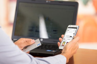 Closeup of young mans hands holding smartphone up with Amazone website visible, Visa card in other hand with laptop computer sitting on desk