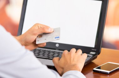 Closeup of man sitting by desk with laptop computer holding up Visa credit card in front of screen as in shopping online