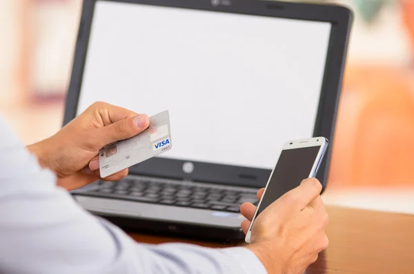 Closeup of young mans hands holding smartphone up, Visa card in other hand with laptop computer sitting on desk — Stockfoto