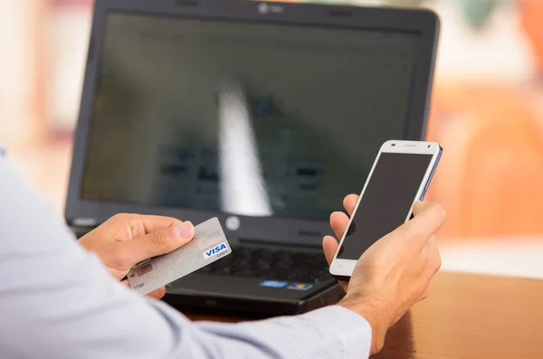 Nahaufnahme junger Männer, die mit der einen Hand das Smartphone hochhalten, mit der anderen die Visakarte, auf deren Schreibtisch ein Laptop sitzt — Stockfoto