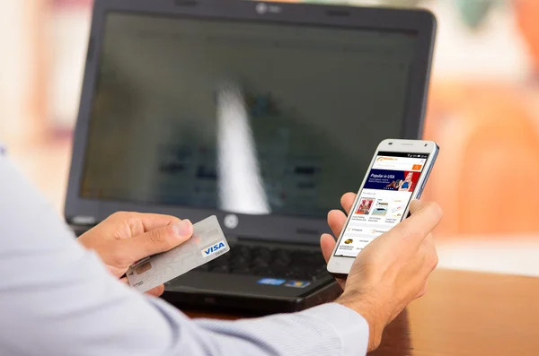 Closeup of young mans hands holding smartphone up with Alibaba website visible, Visa card in other hand with laptop computer sitting on desk — Stock Photo, Image