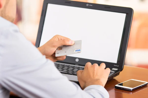 Closeup of man sitting by desk with laptop computer holding up Visa credit card in front of screen as in shopping online — стокове фото