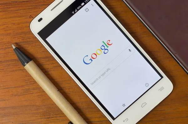 QUITO, ECUADOR - AUGUST 3, 2015: White smartphone lying on wooden desk with screen activated displaying Google search site next to a pen, business and communication concept — 图库照片