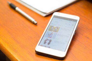 QUITO, ECUADOR - AUGUST 3, 2015: White smartphone lying on wooden desk with unknown website screen next to a pen and notepad, business communication concept