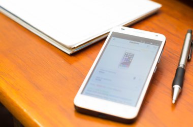 QUITO, ECUADOR - AUGUST 3, 2015: White smartphone lying on wooden desk with Amazon website screen next to a pen and notepad, business communication concept