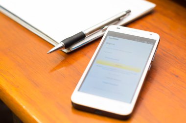 QUITO, ECUADOR - AUGUST 3, 2015: White smartphone lying on wooden desk with Amazon website screen next to a pen and notepad, business communication concept