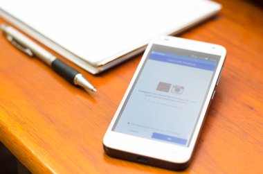 QUITO, ECUADOR - AUGUST 3, 2015: White smartphone lying on wooden desk with Facebook website screen next to a pen and notepad, business communication concept