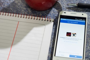 QUITO, ECUADOR - AUGUST 3, 2015: White smartphone lying on table with Facebook website screen open next to a pen, notepad and coffee mug, business communication concept