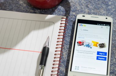 QUITO, ECUADOR - AUGUST 3, 2015: White smartphone lying on table with Ebay website screen open next to a pen, notepad and coffee mug, business communication concept