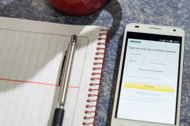 QUITO, ECUADOR - AUGUST 3, 2015: White smartphone lying on table with Amazon website screen open next to a pen, notepad and coffee mug, business communication concept