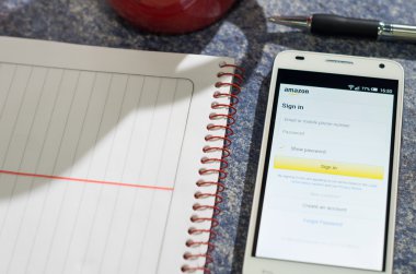 QUITO, ECUADOR - AUGUST 3, 2015: White smartphone lying on table with Amazon website screen open next to a pen, notepad and coffee mug, business communication concept