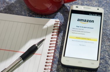 QUITO, ECUADOR - AUGUST 3, 2015: White smartphone lying on table with Amazon website screen open next to a pen, notepad and coffee mug, business communication concept