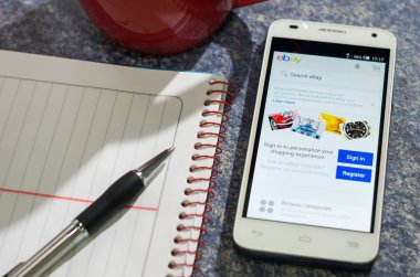 QUITO, ECUADOR - AUGUST 3, 2015: White smartphone lying on table with Ebay website screen open next to a pen, notepad and coffee mug, business communication concept