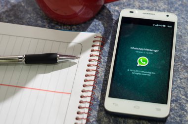 QUITO, ECUADOR - AUGUST 3, 2015: White smartphone lying on table with WhatsApp website screen open next to a pen, notepad and coffee mug, business communication concept