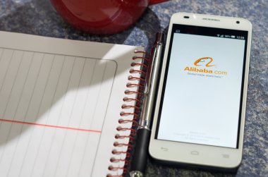QUITO, ECUADOR - AUGUST 3, 2015: White smartphone lying on table with Alibaba website screen open next to a pen, notepad and coffee mug, business communication concept