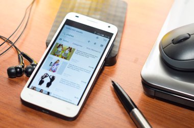 QUITO, ECUADOR - AUGUST 3, 2015: White smartphone lying on wooden desk with Amazon website screen next to a pen, mouse and laptop, business communication concept