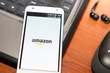 QUITO, ECUADOR - AUGUST 3, 2015: White smartphone closeup lying on laptop corner with Amazon website screen visible, eaplugs mouse and pen blurry background, business communication concept
