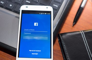 QUITO, ECUADOR - AUGUST 3, 2015: White smartphone closeup lying on laptop corner with Facebook website screen visible, eaplugs mouse and pen blurry background, business communication concept