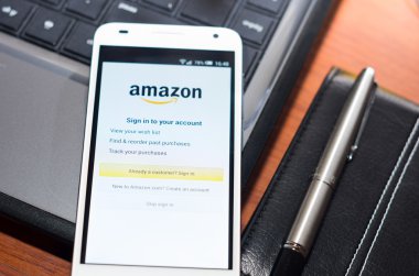 QUITO, ECUADOR - AUGUST 3, 2015: White smartphone closeup lying on laptop corner with Amazon website screen visible, eaplugs mouse and pen blurry background, business communication concept