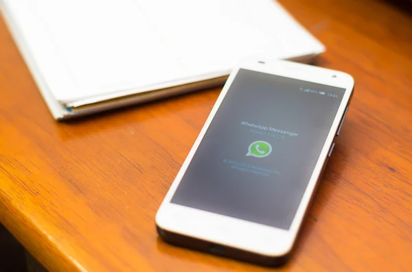 QUITO, ECUADOR - AUGUST 3, 2015: White smartphone lying on wooden desk with WhatsApp screen open next to a pen and notepad, business communication concept — Stock Fotó