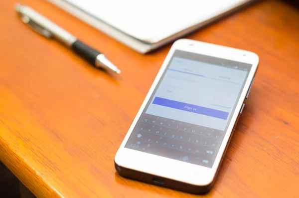 QUITO, ECUADOR - AUGUST 3, 2015: White smartphone lying on wooden desk with unknown login screen open next to a pen and notepad, business communication concept — Stockfoto