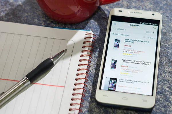 QUITO, ECUADOR - AUGUST 3, 2015: White smartphone lying on table with Amazon website screen open next to a pen, notepad and coffee mug, business communication concept — Stockfoto