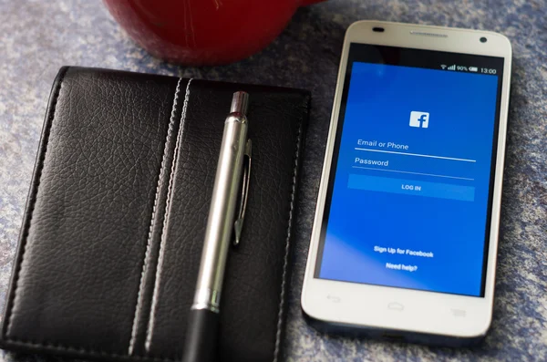 QUITO, ECUADOR - AUGUST 3, 2015: White smartphone lying on desk with Facebook website screen next to a pen and notepad, business communication concept — Stok fotoğraf