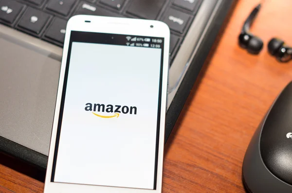 QUITO, ECUADOR - AUGUST 3, 2015: White smartphone closeup lying on laptop corner with Amazon website screen visible, eaplugs mouse and pen blurry background, business communication concept — Stock Photo, Image