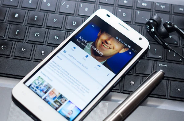 QUITO, ECUADOR - AUGUST 3, 2015: White smartphone closeup lying next to silver pen on laptop keyboard with Mark Zuckerberg Facebook profile screen visible — 图库照片