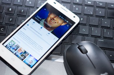 QUITO, ECUADOR - AUGUST 3, 2015: White smartphone closeup lying next to silver pen on laptop keyboard with Mark Zuckerberg Facebook profile screen visible