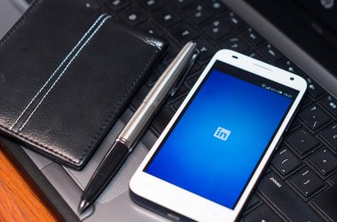 QUITO, ECUADOR - AUGUST 3, 2015: White smartphone closeup lying next to silver pen and wallet on laptop keyboard with in website logo screen visible