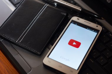 QUITO, ECUADOR - AUGUST 3, 2015: White smartphone closeup lying next to silver pen and wallet on laptop keyboard with YouTube play symbol visible screen