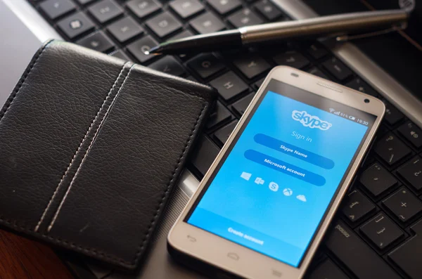 QUITO, ECUADOR - AUGUST 3, 2015: White smartphone closeup lying next to silver pen and wallet on laptop keyboard with Skype login screen visible