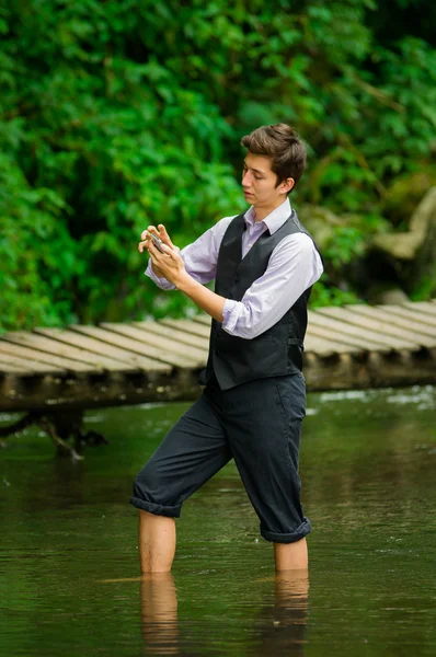 Młody mężczyzna elegancki przy użyciu telefonu komórkowego w lesie nad jeziorem — Zdjęcie stockowe