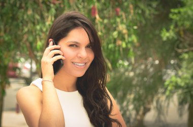 Classy attractive brunette wearing white dress talking on the phone in outdoors environment