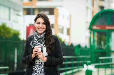 Brunette model wearing black jacket and grey scarf waiting for public transportation acting cold at station with cup of coffee