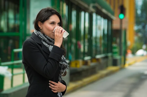 Morena modelo vestindo casaco preto e cachecol cinza à espera de transporte público agindo frio na estação com xícara de café — Fotografia de Stock