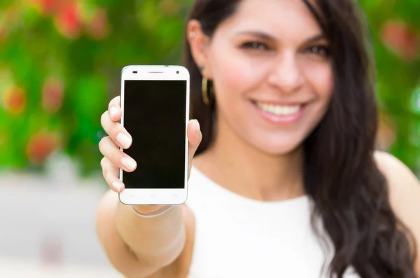 Classy attractive brunette wearing white dress holding up mobile phone in outdoors environment smiling — Stock Photo, Image