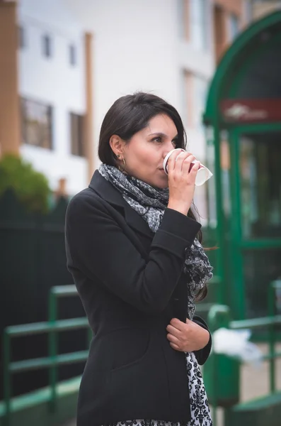 Brunette model wearing black jacket and grey scarf waiting for public transportation acting cold at station with cup of coffee — Stok fotoğraf