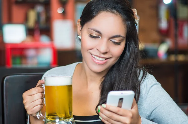 Modelo morena sentado à mesa do restaurante segurando copo de cerveja e usando telefone celular enquanto sorri — Fotografia de Stock