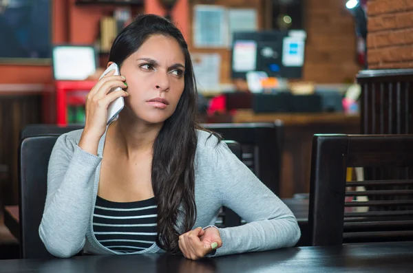 Attractive brunette wearing grey sweater sitting at restaurant table talking on mobile phone with worried facial expression — Stock Photo, Image