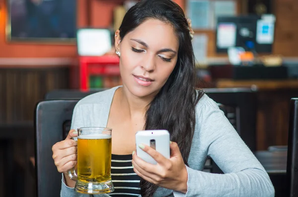 Esmer modeli bardak bira tutarak ve gülümseyen iken cep telefonunuzu kullanarak Restoran tablo başına oturmuş — Stok fotoğraf