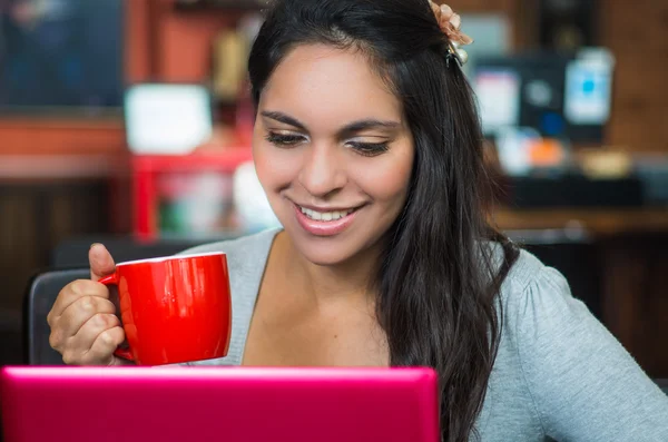 Attraktive Brünette mit grauem Pullover sitzt am Restauranttisch und arbeitet mit rosa Laptop und hält roten Kaffeebecher vor dem Mund — Stockfoto