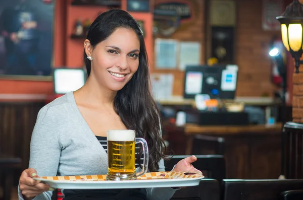 Brunette model waitress at restaurant smiling happily carrying a tray with glass fo beer on it — Stock Photo, Image